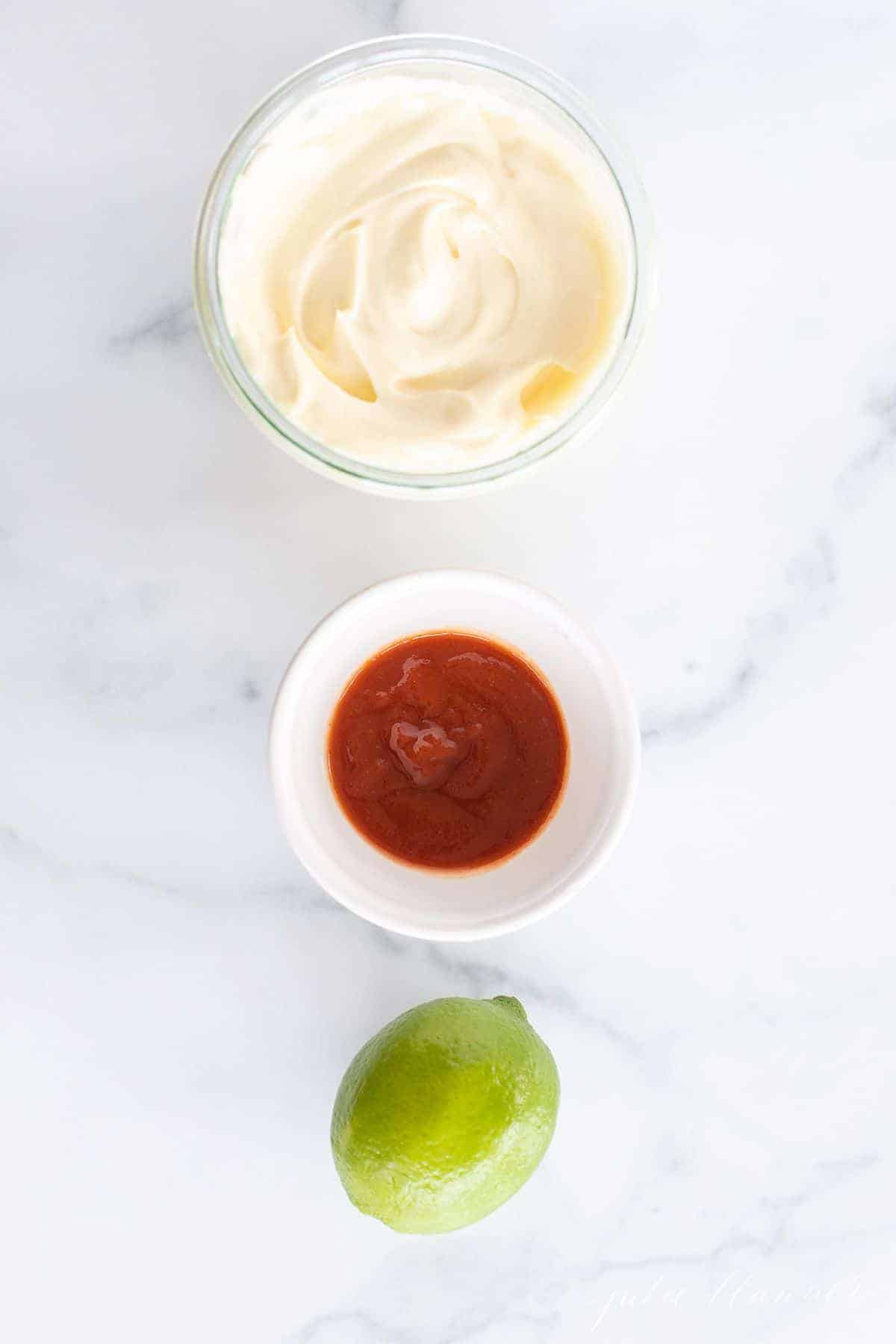 A bowl of mayonnaise, a bowl of sriracha and a whole lime, sitting in a row on a marble surface.