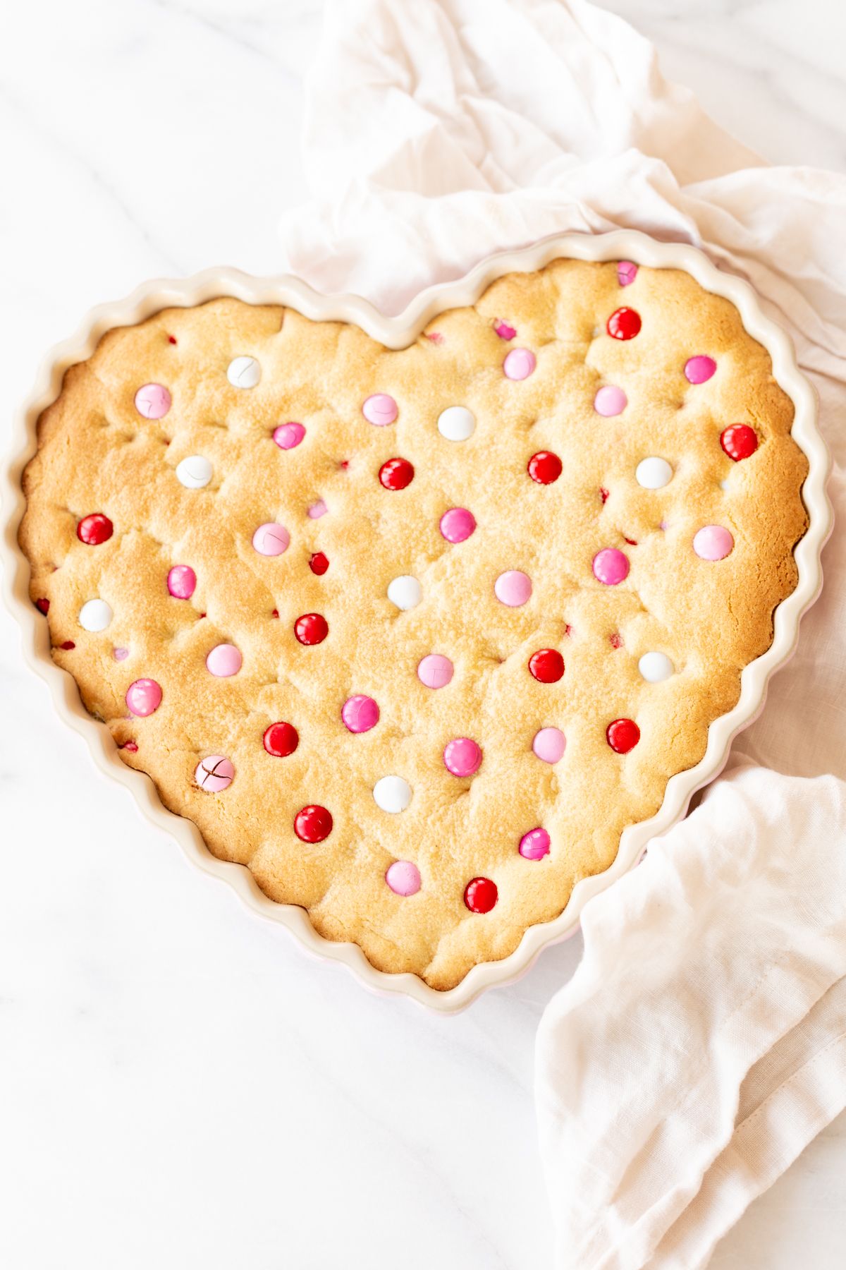 Thick and Chewy M&M Cake in a heart shaped dish, topped with Valentine's Day M&Ms. A linen napkin is to the side.