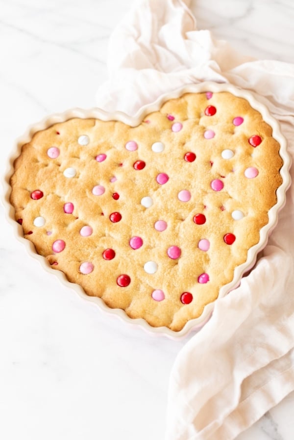 Thick and Chewy M&M Cake in a heart shaped dish, topped with Valentine's Day M&Ms. A linen napkin is to the side.