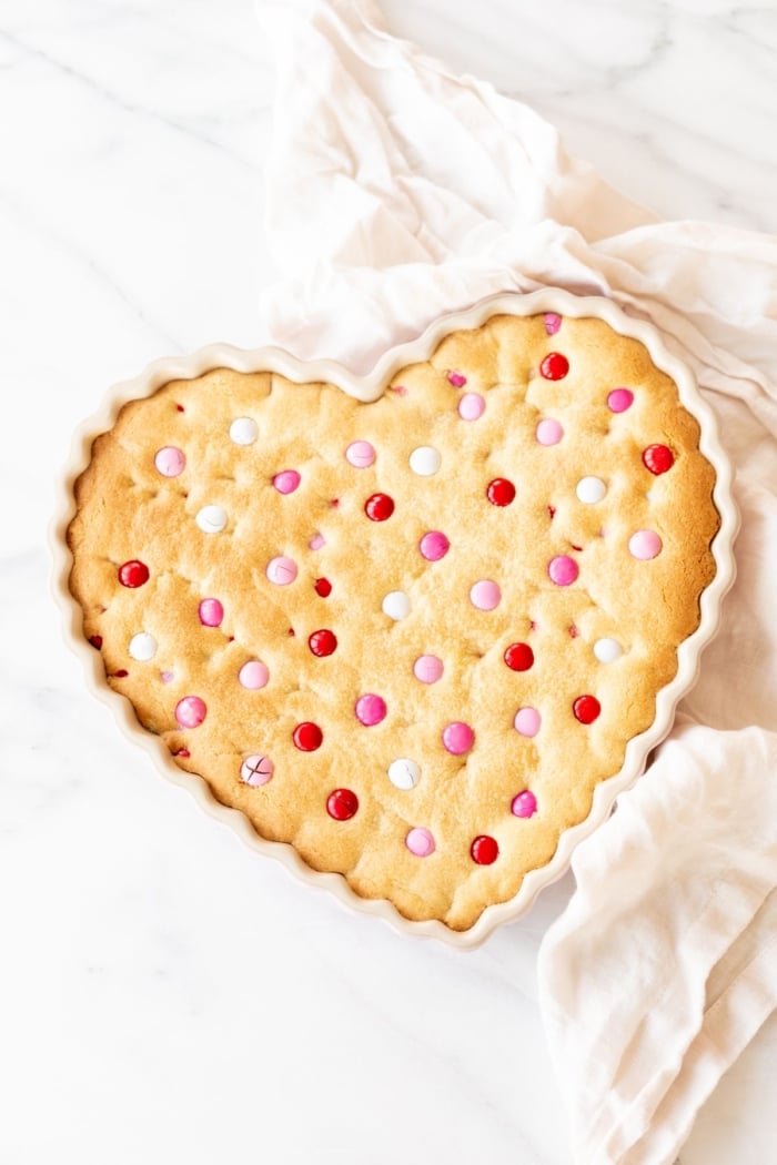 Thick and Chewy M&M Cake in a heart shaped dish, topped with Valentine's Day M&Ms. A linen napkin is to the side.