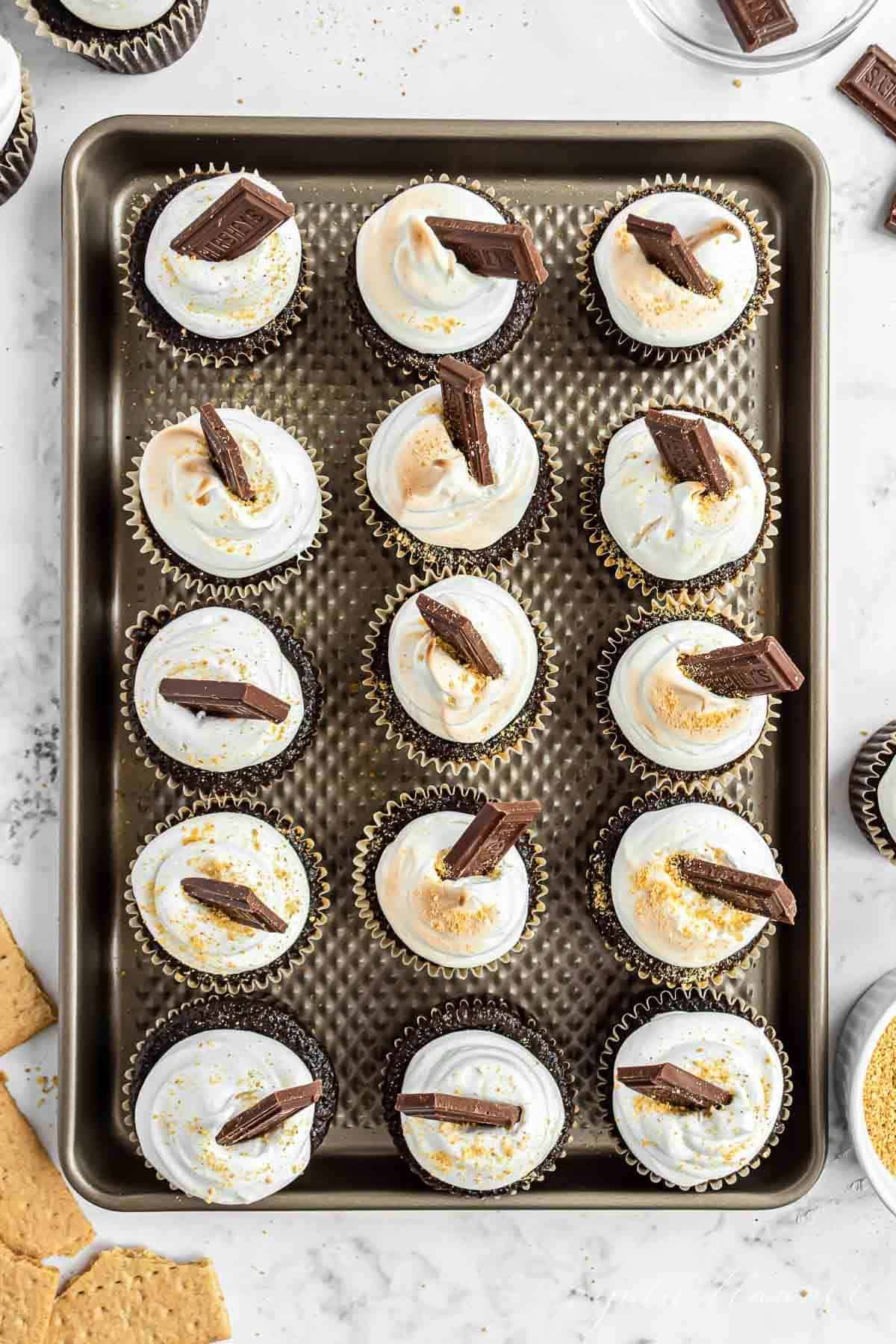 Homemade smores cupcakes on a baking sheet, lined up and topped with marshmallow frosting and a piece of Hersheys bar.