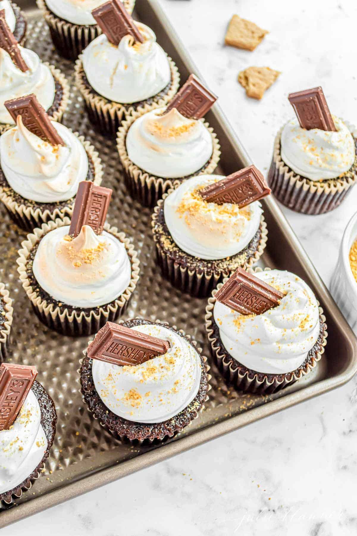 Homemade smores cupcakes on a baking sheet, lined up and topped with marshmallow frosting and a piece of Hersheys bar.