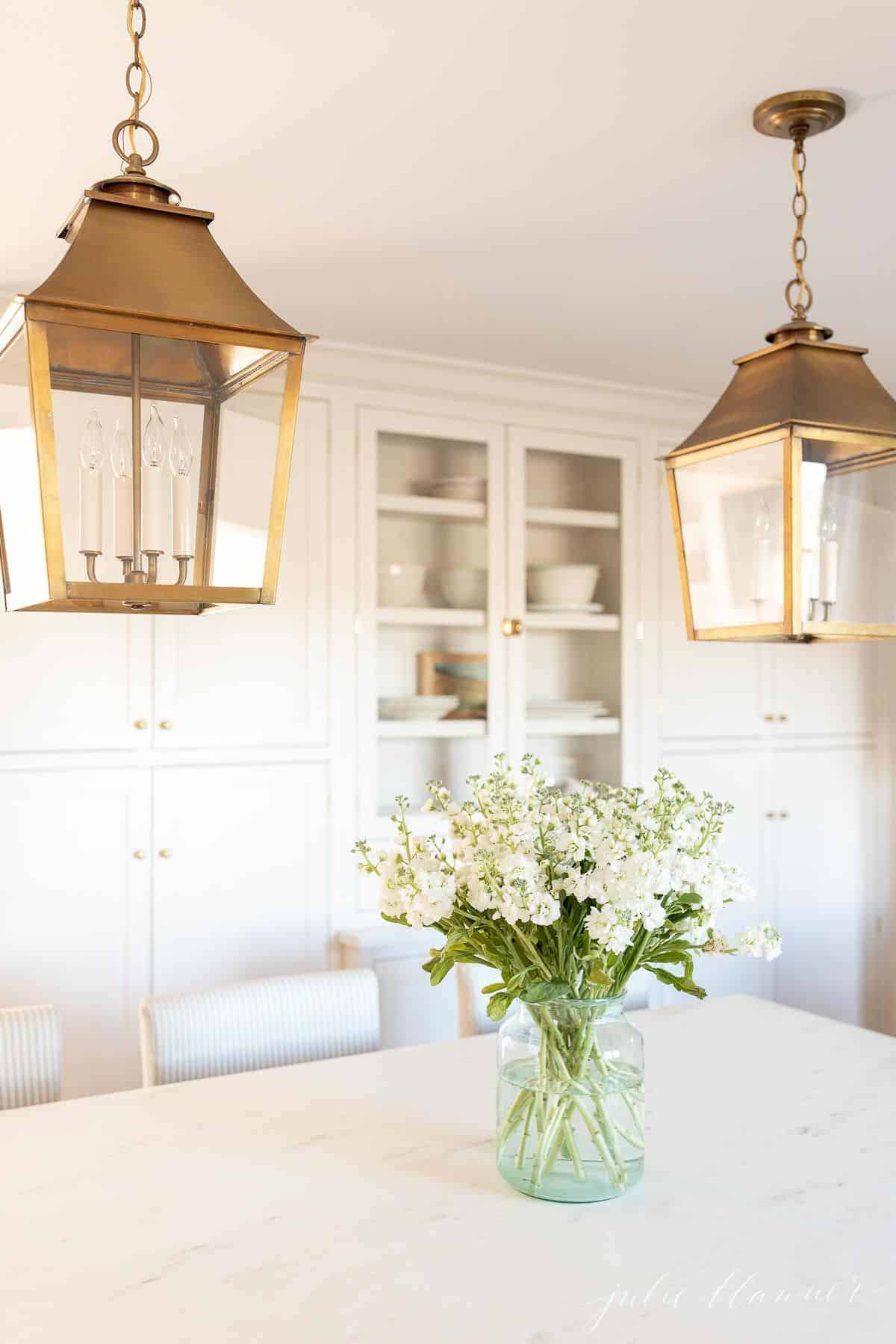 A white kitchen with brass lanterns and marble countertops, with a stock flower arrangement on the island.