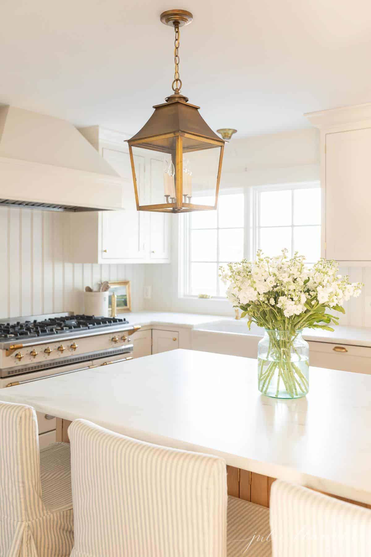 A white kitchen with brass lanterns and marble countertops, with a stock flower arrangement on the island.