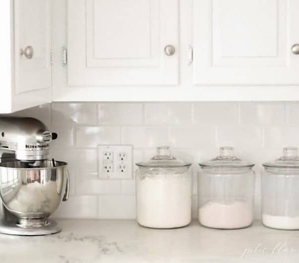 A white kitchen with marble countertops, a stand mixer and glass canisters of baking supplies.