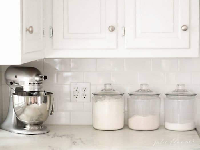 A white kitchen with marble countertops, a stand mixer and glass canisters of baking supplies.