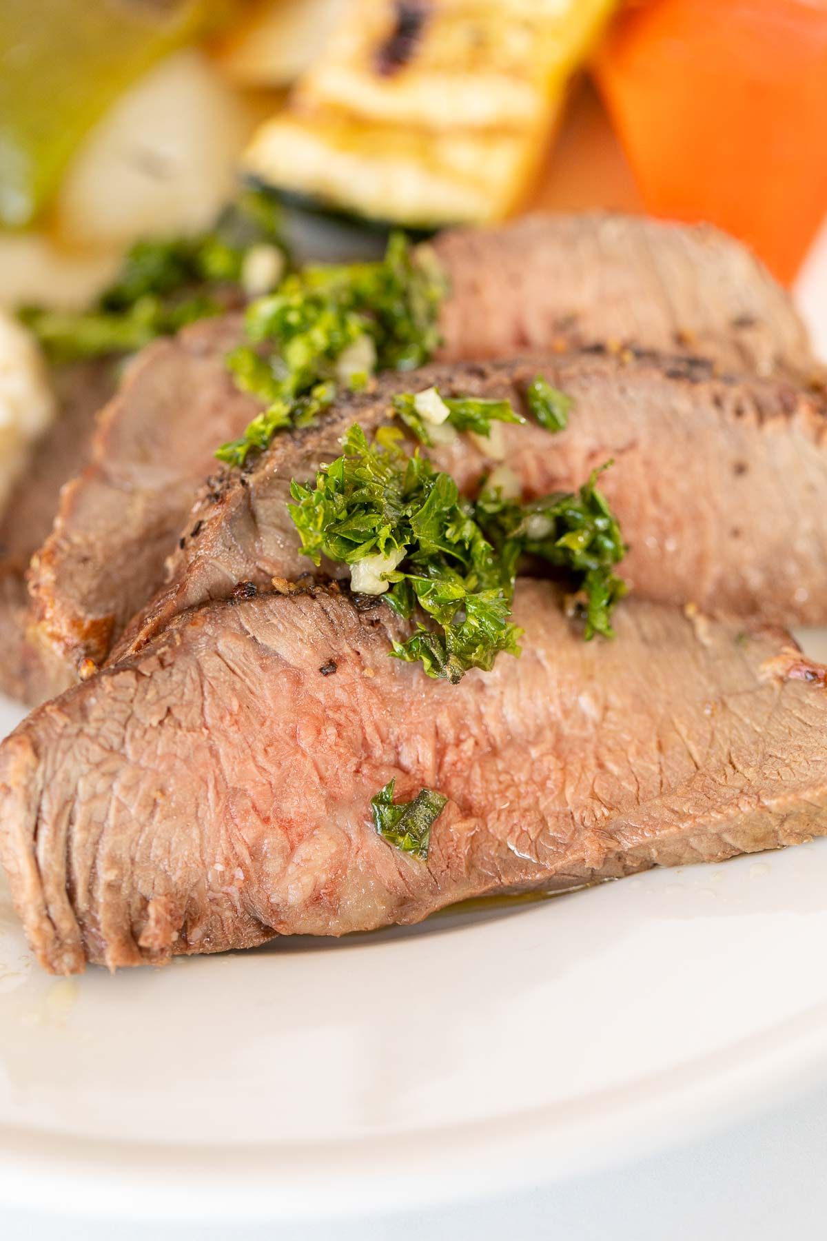 Sliced broiled steak topped with chopped herbs and garlic on a white plate, with grilled vegetables in the background. A perfect example of a broiled steak recipe that showcases how to broil steak to perfection in the oven.