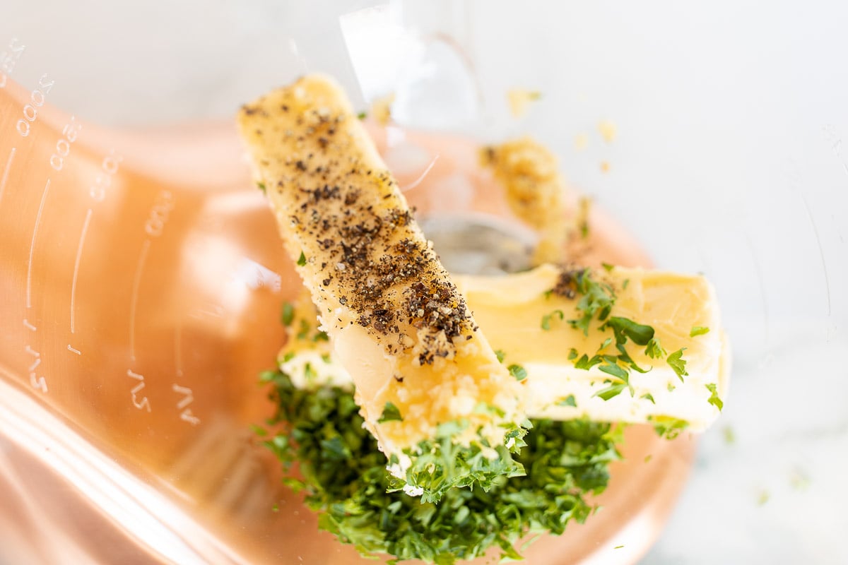 A stick of butter and fresh herbs inside the glass bowl of a mixer.