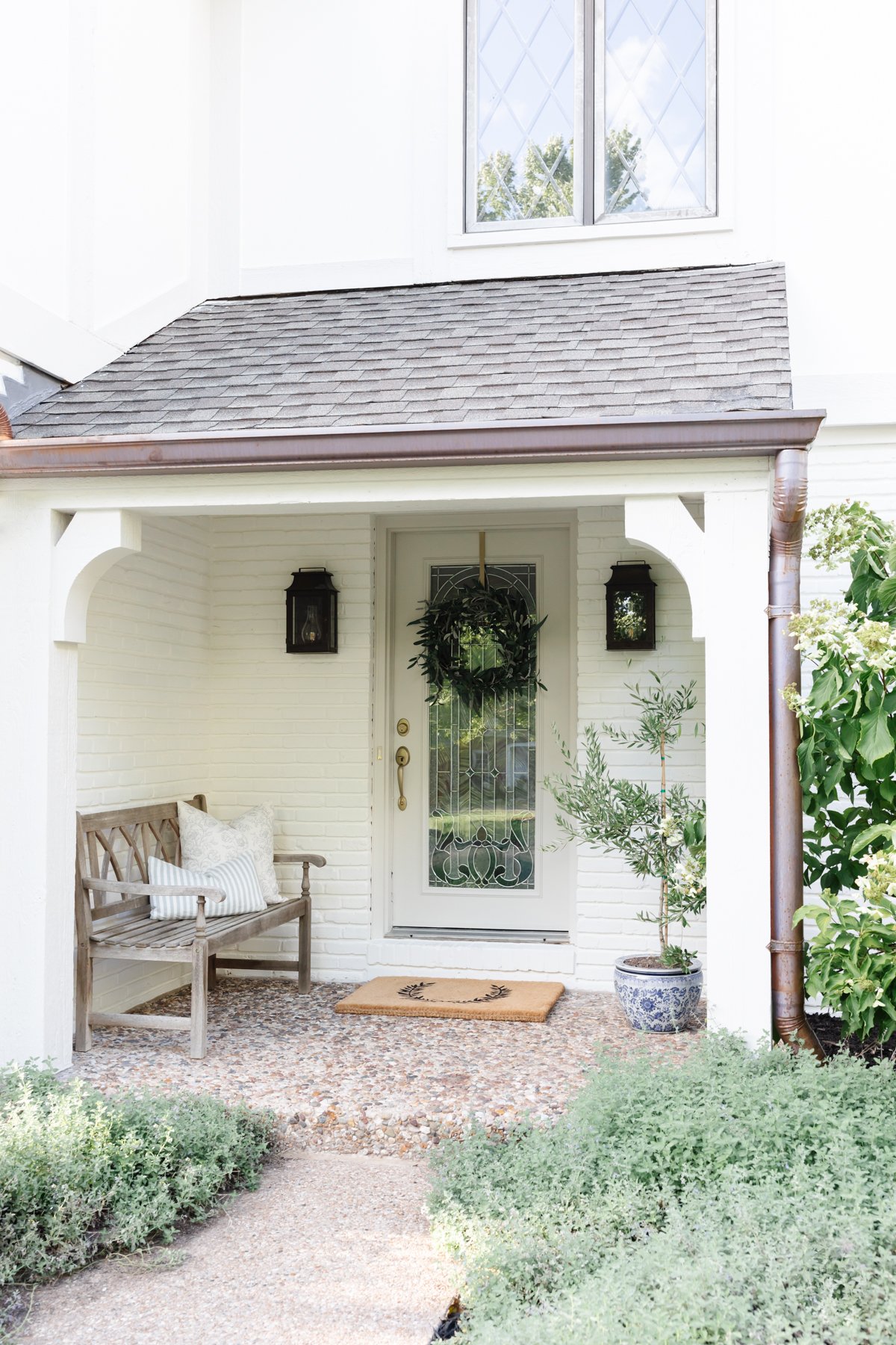 A white front porch adorned with an olive tree, bench, and plants.