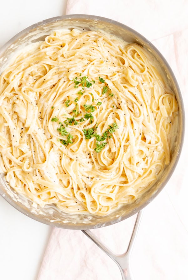 A skillet filled with creamy fettuccine Alfredo, crafted from a classic fettuccine Alfredo recipe, garnished with chopped parsley and cracked black pepper.