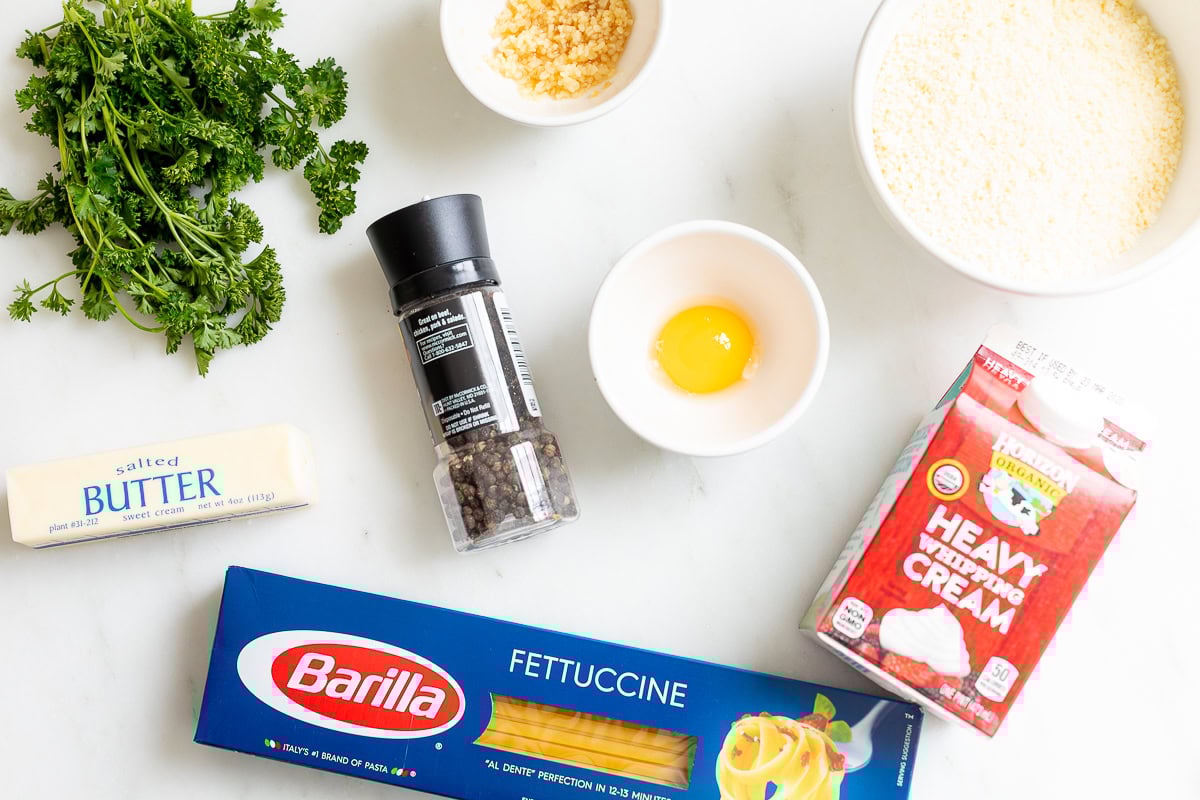 A delightful fettuccine alfredo spread awaits, with ingredients on the table: parsley, butter, black pepper, an egg, grated cheese, and heavy whipping cream alongside a box of fettuccine. Perfect for crafting a rich alfredo sauce recipe that promises indulgence in every bite.
