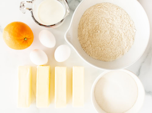 Ingredients for baking orange pound cake arranged on a white surface, including flour, sugar, butter, eggs, milk, and an orange.