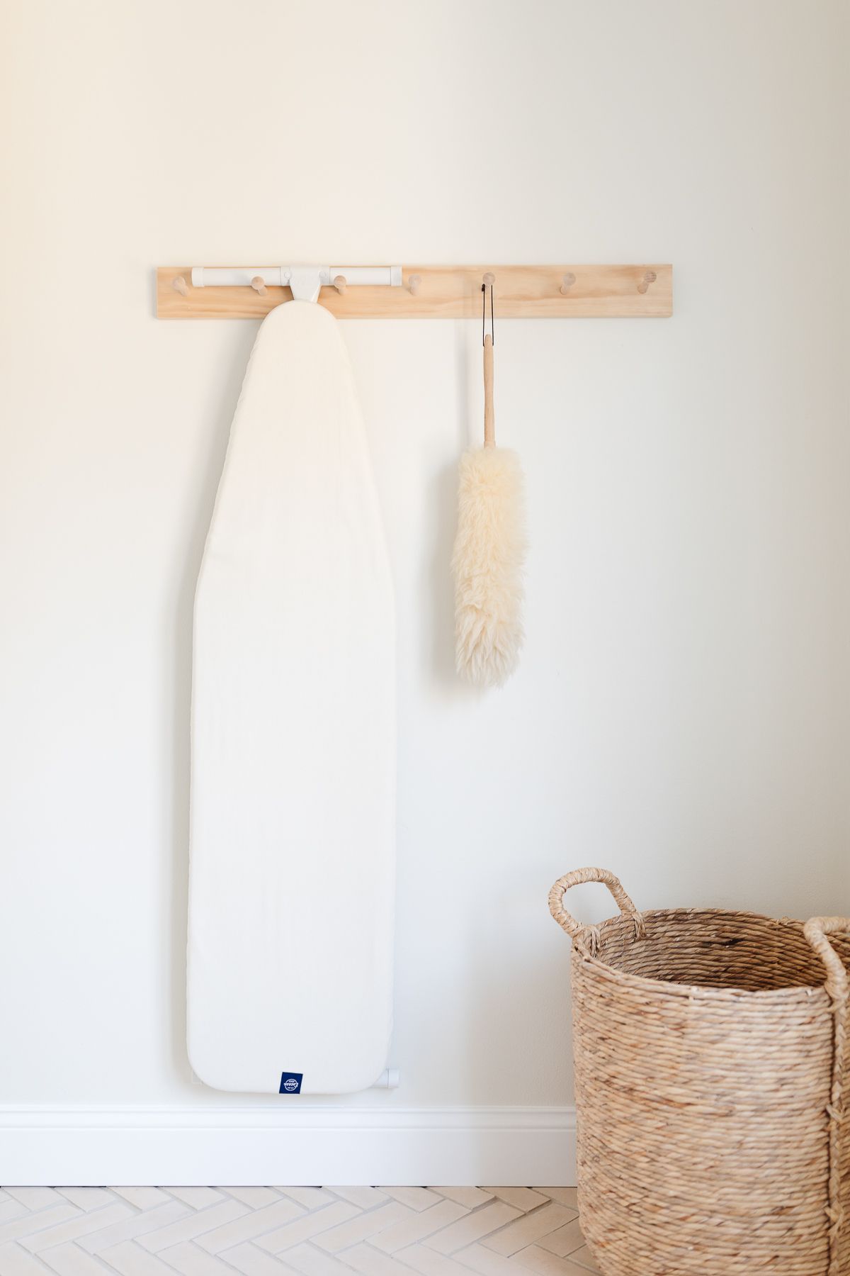 A peg rail on a white wall holding an ironing board and feather duster.