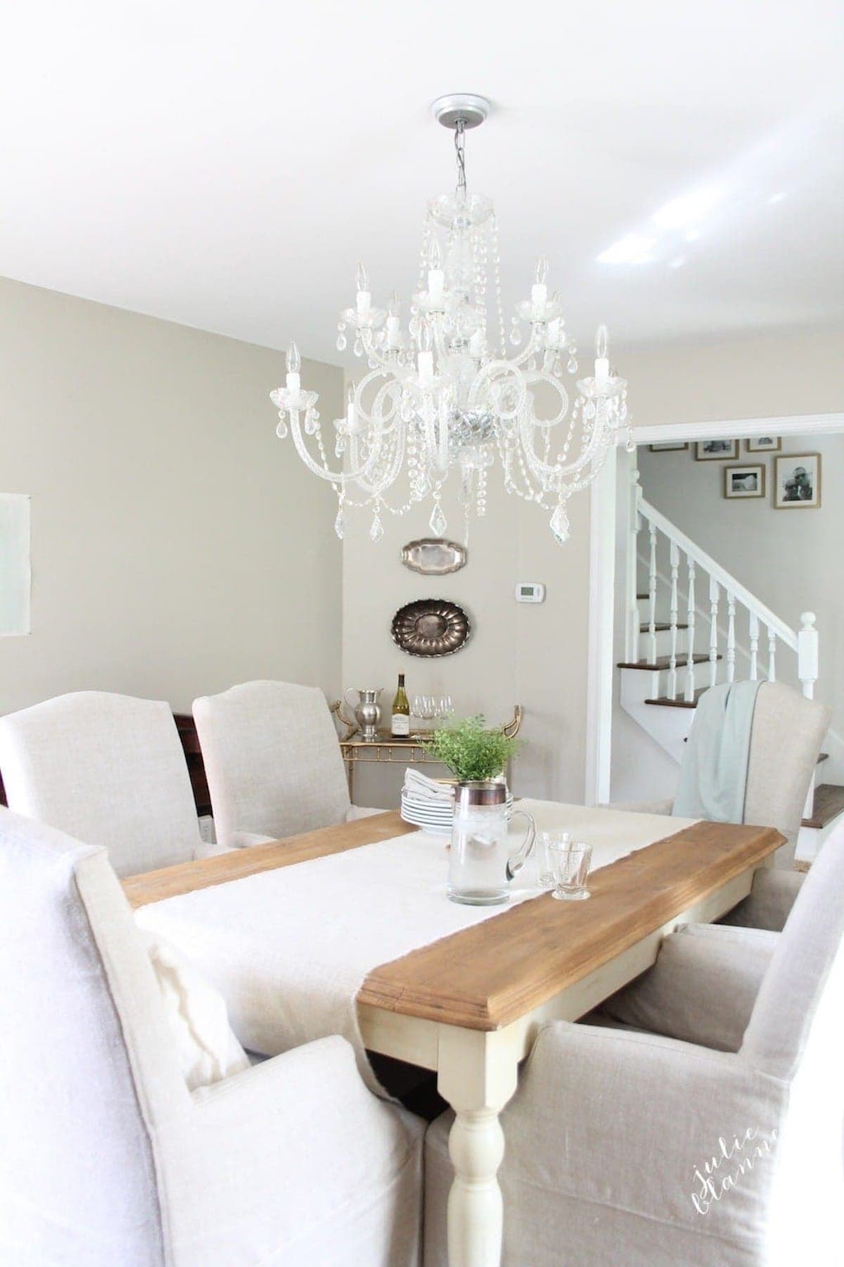 A dining room with a wooden table, beige upholstered chairs in Sherwin Williams Accessible Beige, a crystal chandelier, and minimalist decor. In the background, there is a staircase and some framed photos on the wall.