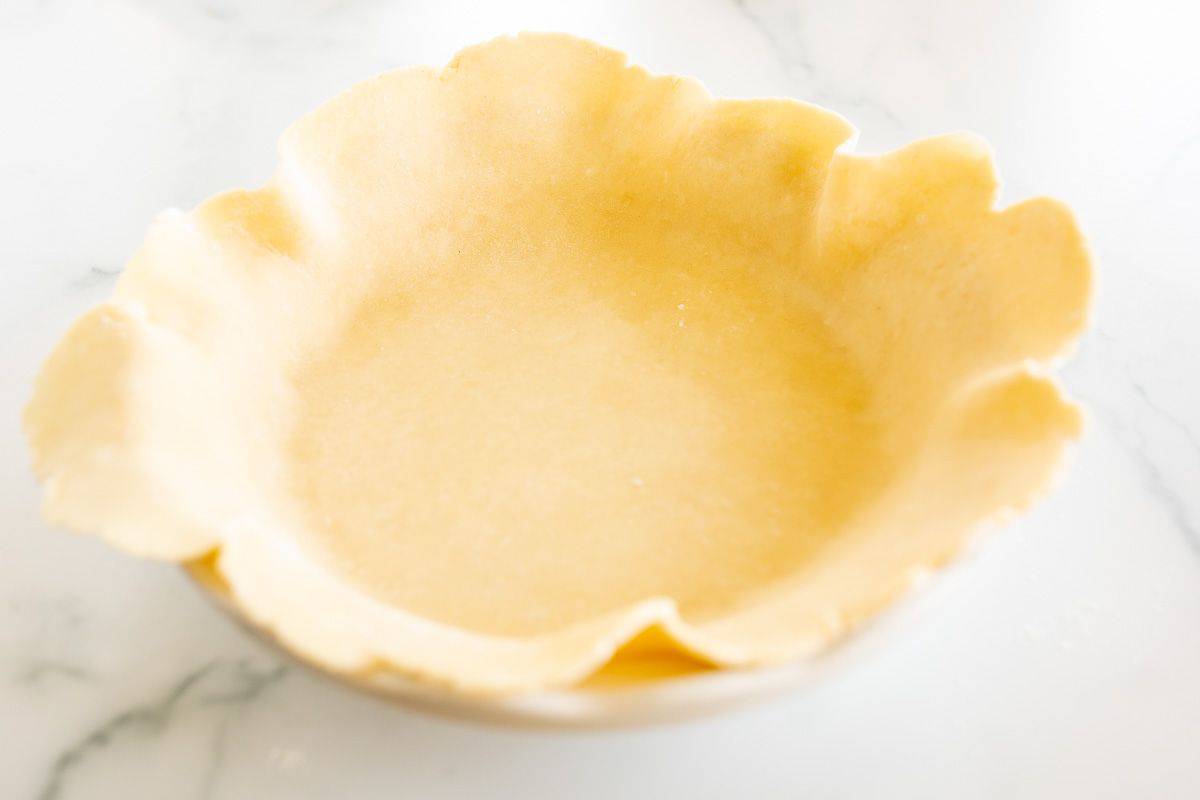 A homemade quiche crust in a fluted pie pan on a white marble surface.