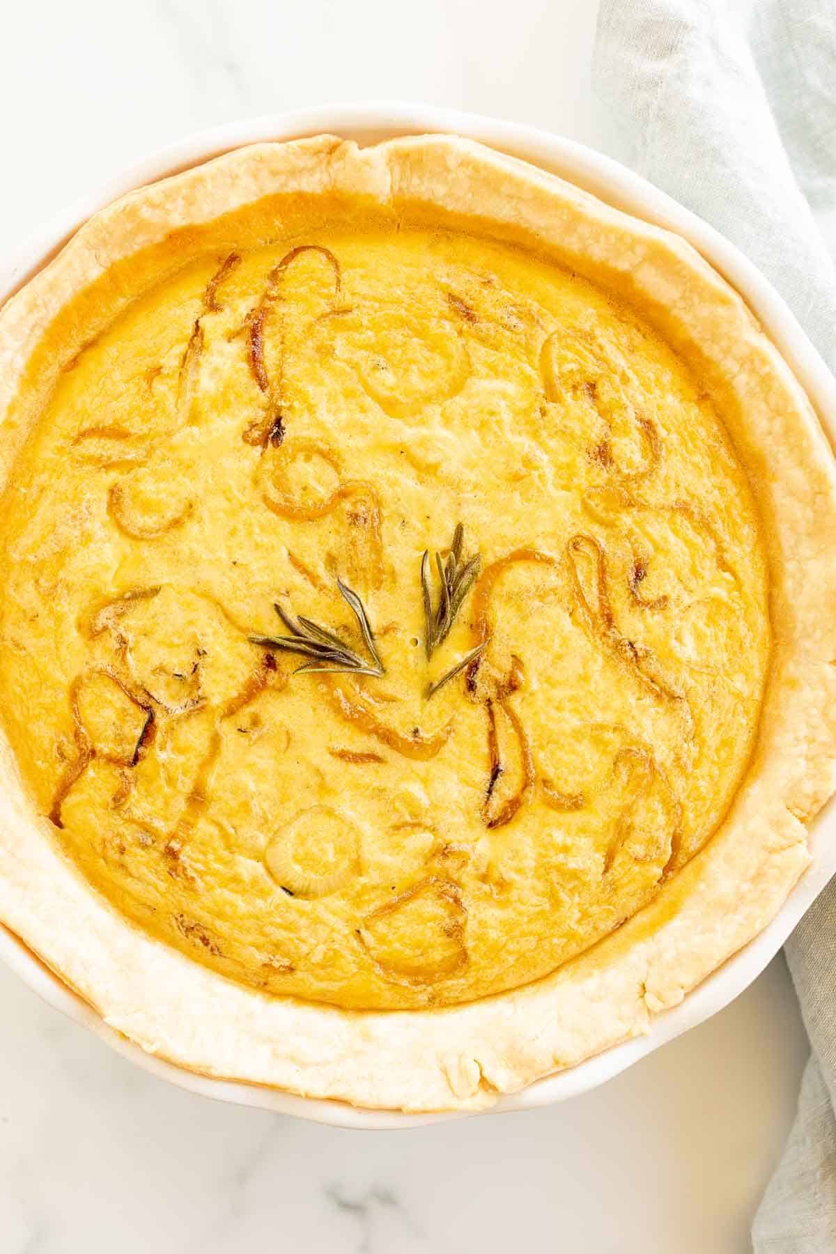 A fully baked quiche in a white pie dish on a marble surface, topped with fresh herbs.