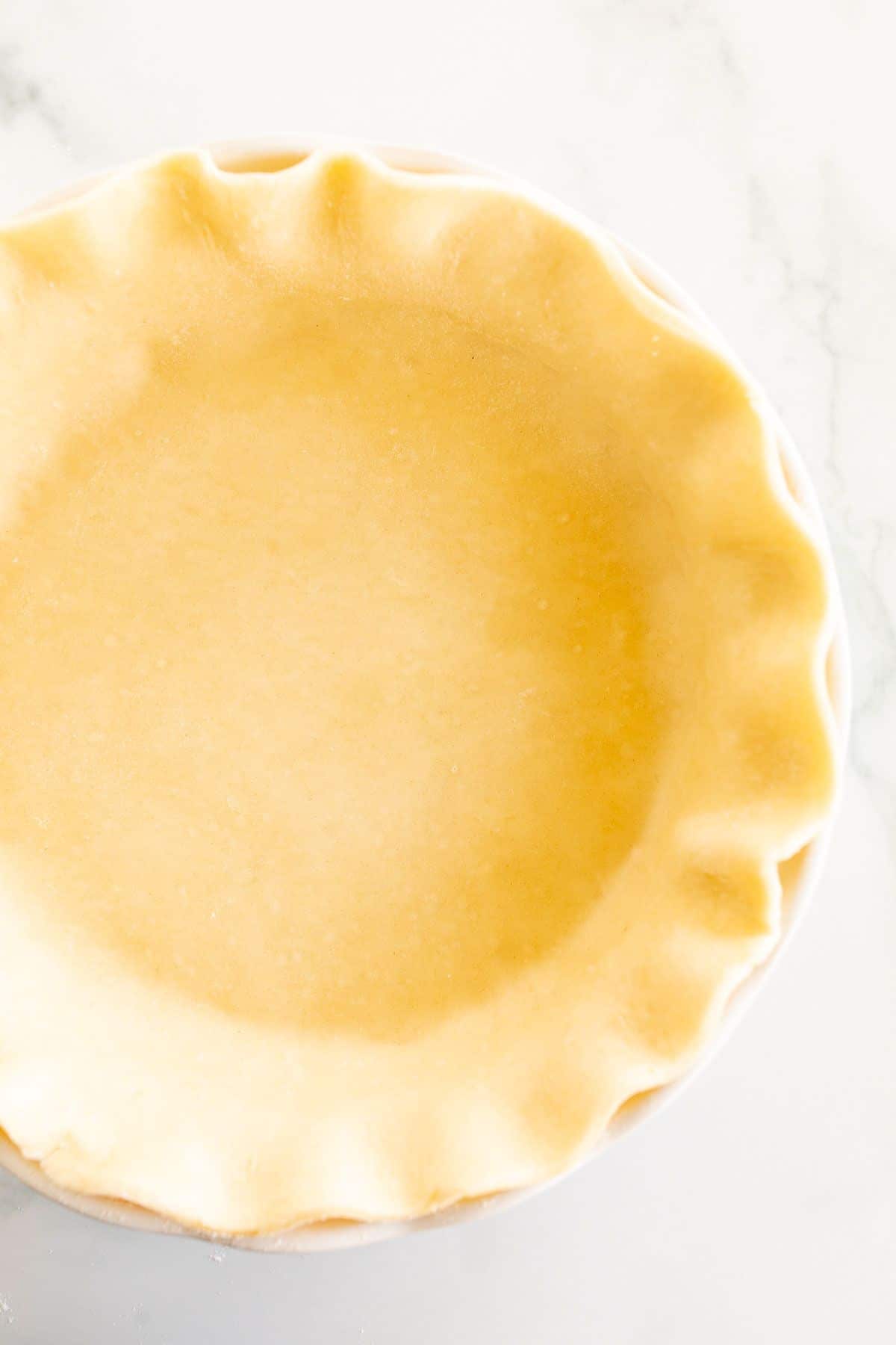 A homemade quiche crust in a fluted pie pan on a white marble surface.