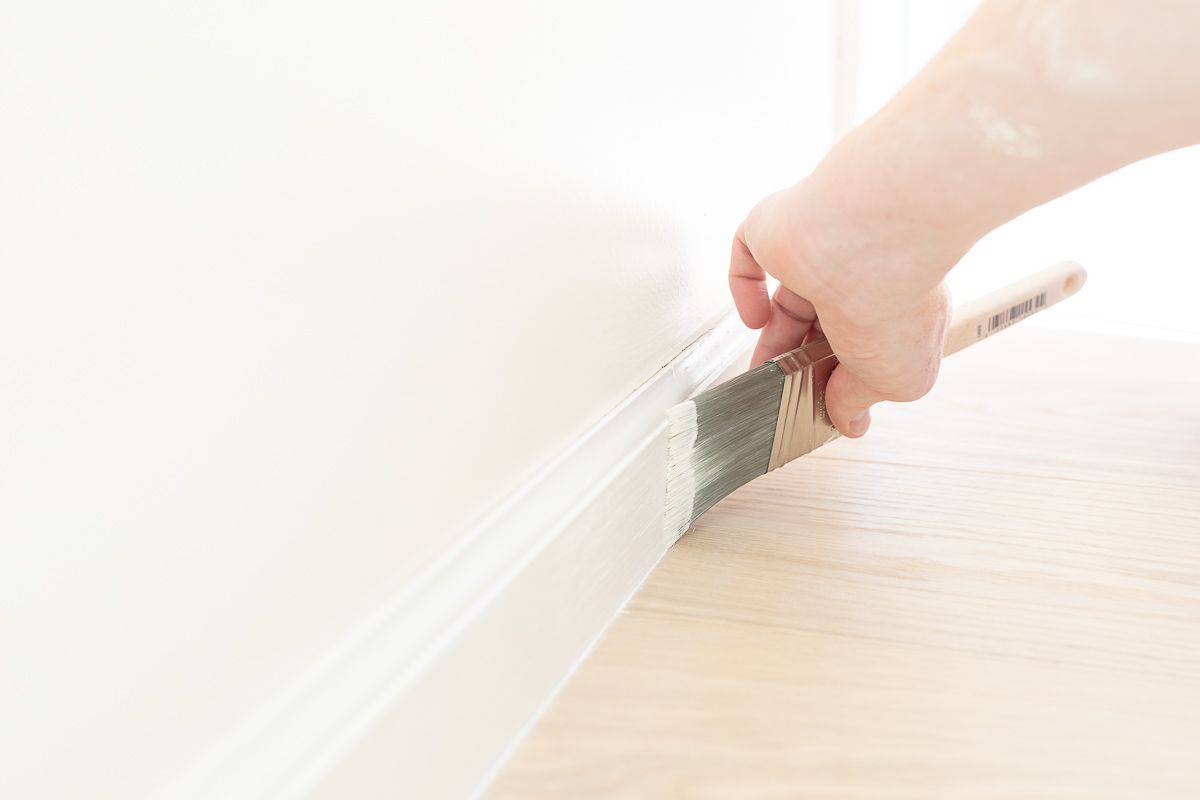 A hand painting trim white with a brush, next to a light wood floor.