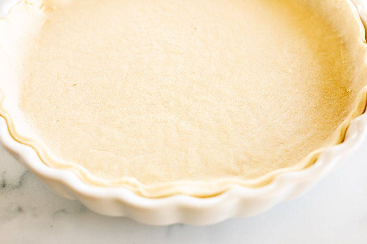 A homemade quiche crust in a fluted pie pan on a white marble surface.