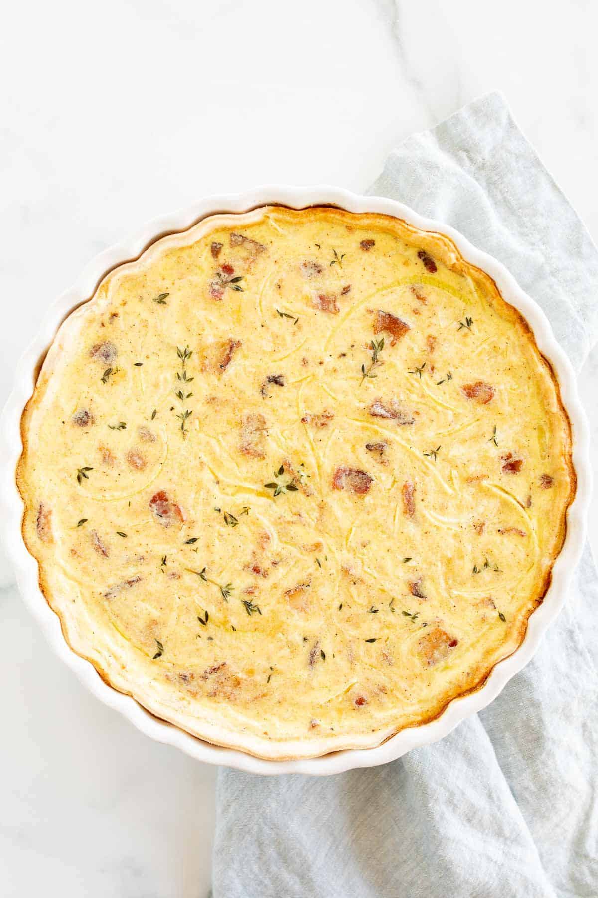 A whole quiche in a white pie dish on a white marble surface.