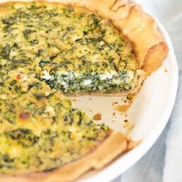 A spinach and feta quiche in a white pie pan, one slice removed.