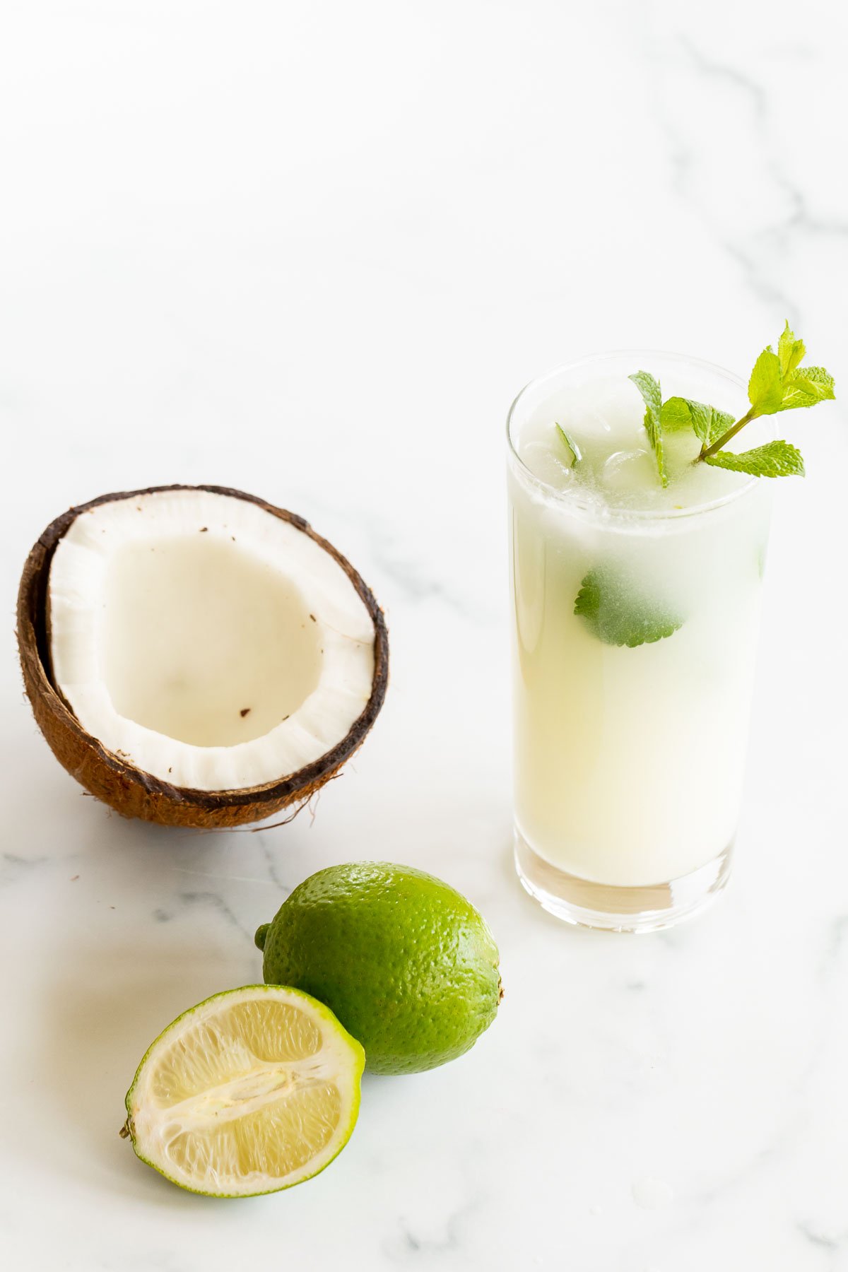 A coconut mojito garnished with mint on a marble surface. Limes and an open coconut to the side.