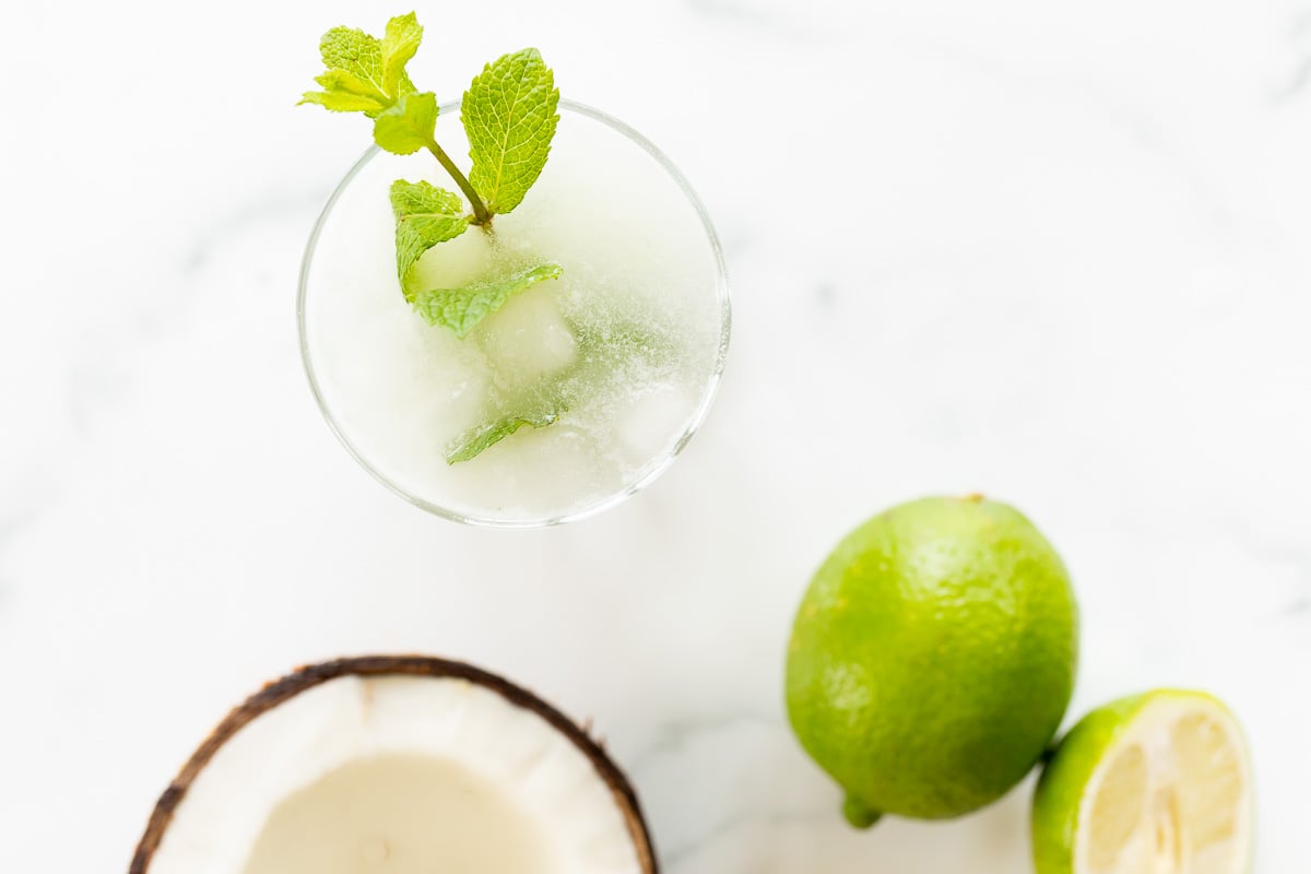 A coconut mojito garnished with mint on a marble surface. Limes and an open coconut to the side.