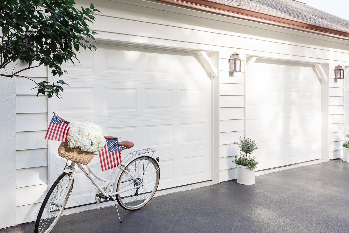 A three car garage on a home painted in a soft white color, with copper lanterns and a bike decorated for the 4th of july