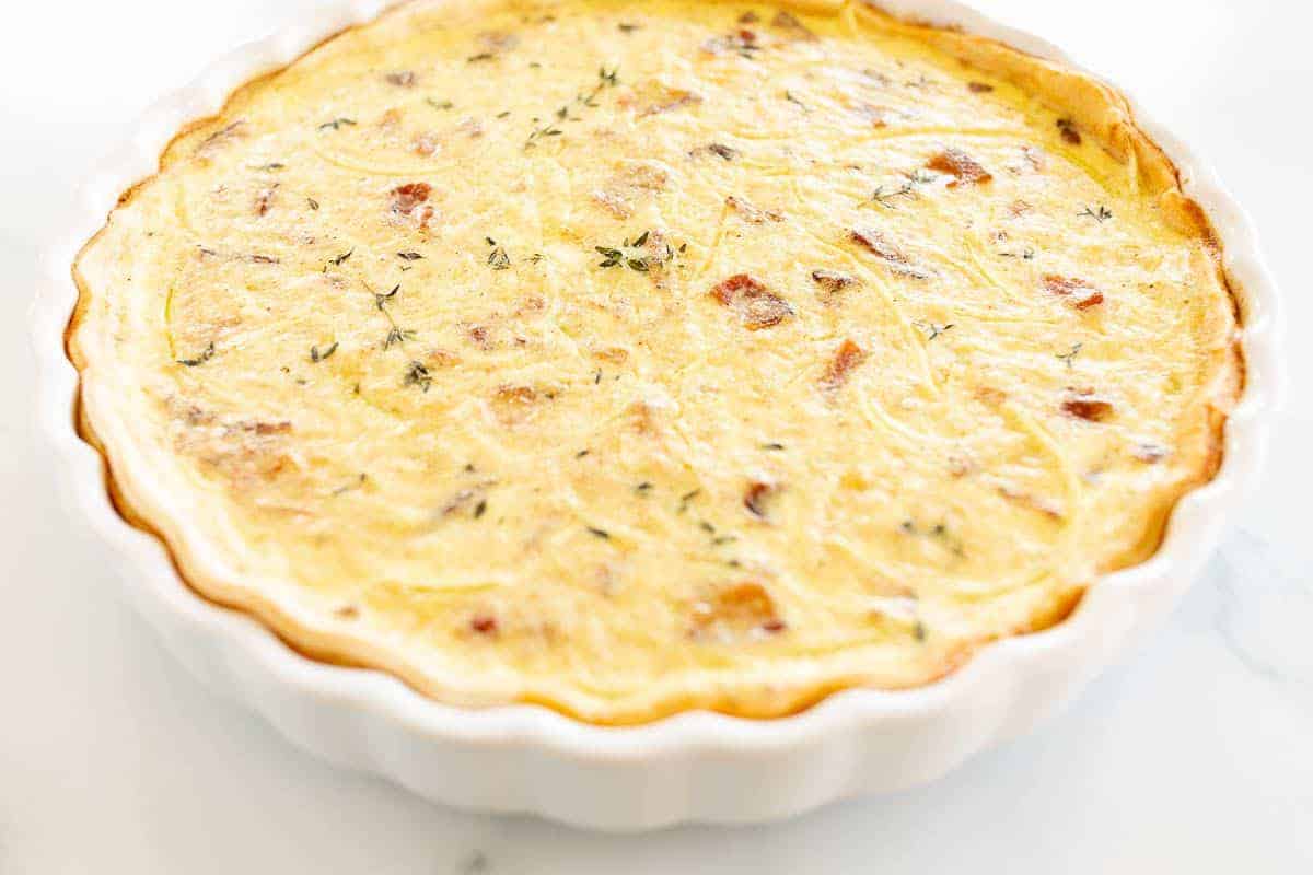 A quiche lorraine baked into a white tart pan, resting on a marble surface.