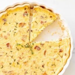 A quiche lorraine recipe in a white pan, one slice removed and another being cut away.