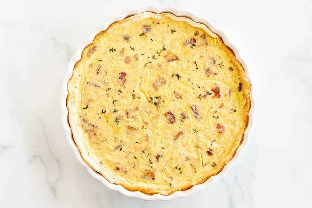 A quiche lorraine baked into a white tart pan, resting on a marble surface.