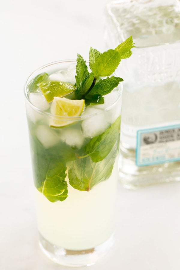 A tequila mojito cocktail with mint leaves and lime in a tall glass, next to a bottle of clear liquor on a white surface.