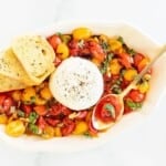 baked burrata bruschetta in a white oval bowl, gold spoon and crostini to the side.