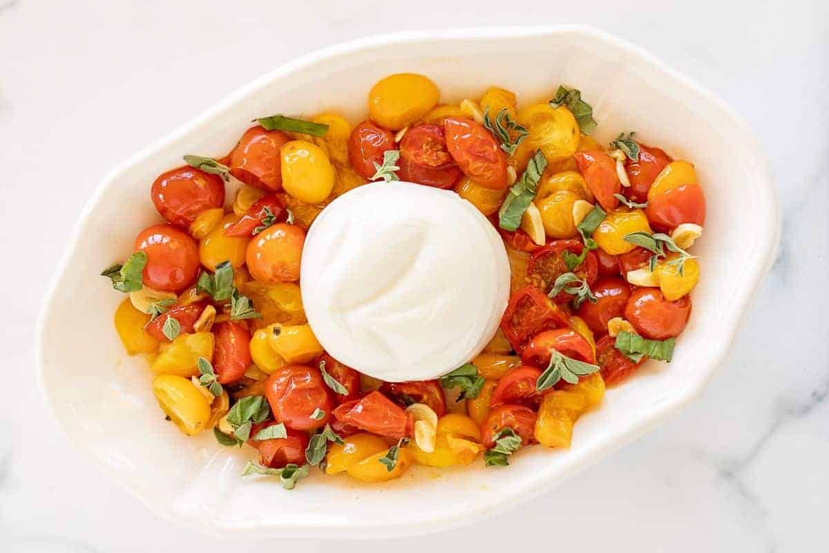 Roasted cherry tomatoes and basil topped with burrata cheese in a white serving dish.