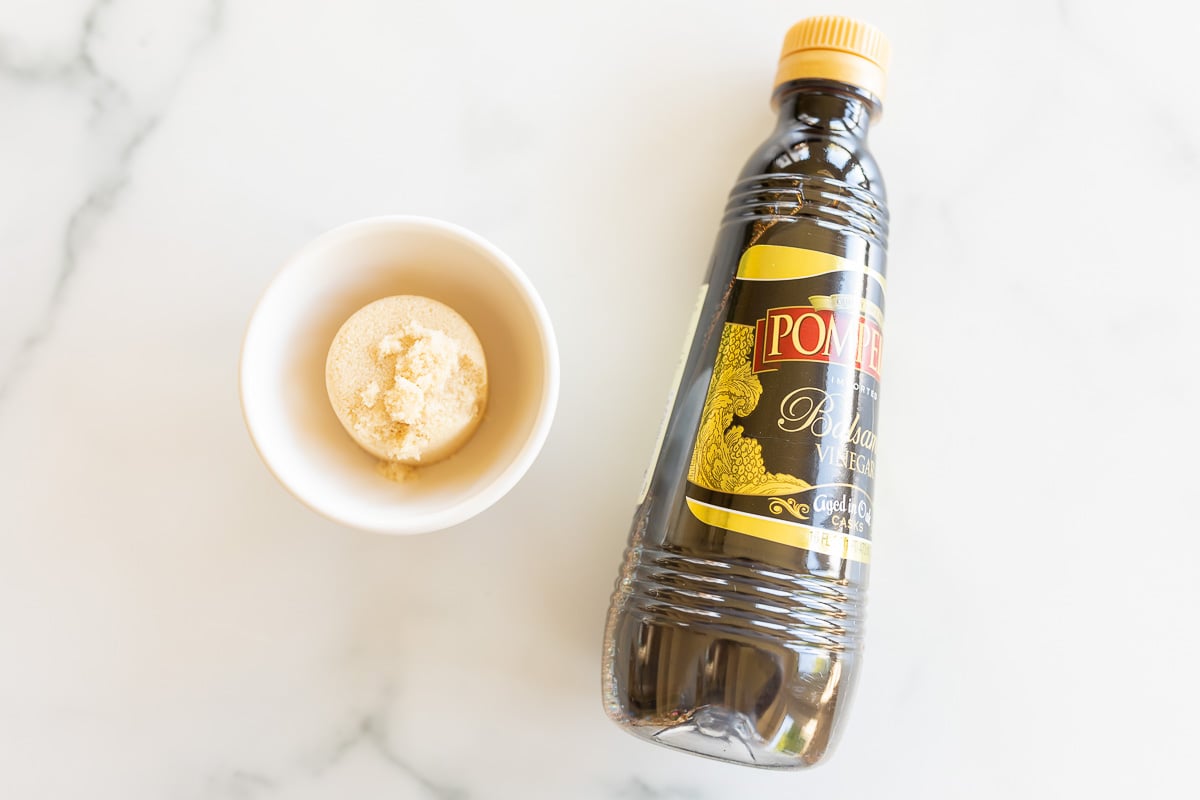 A bottle of balsamic vinegar and a small ramekin with brown sugar on a marble countertop.