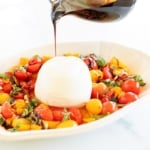 A small glass pitcher of balsamic glaze pouring over a dish of cherry tomatoes and mozzarella.