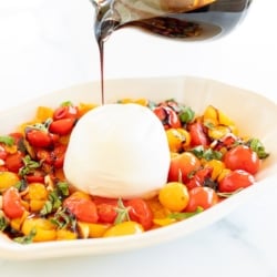 A small glass pitcher of balsamic glaze pouring over a dish of cherry tomatoes and mozzarella.