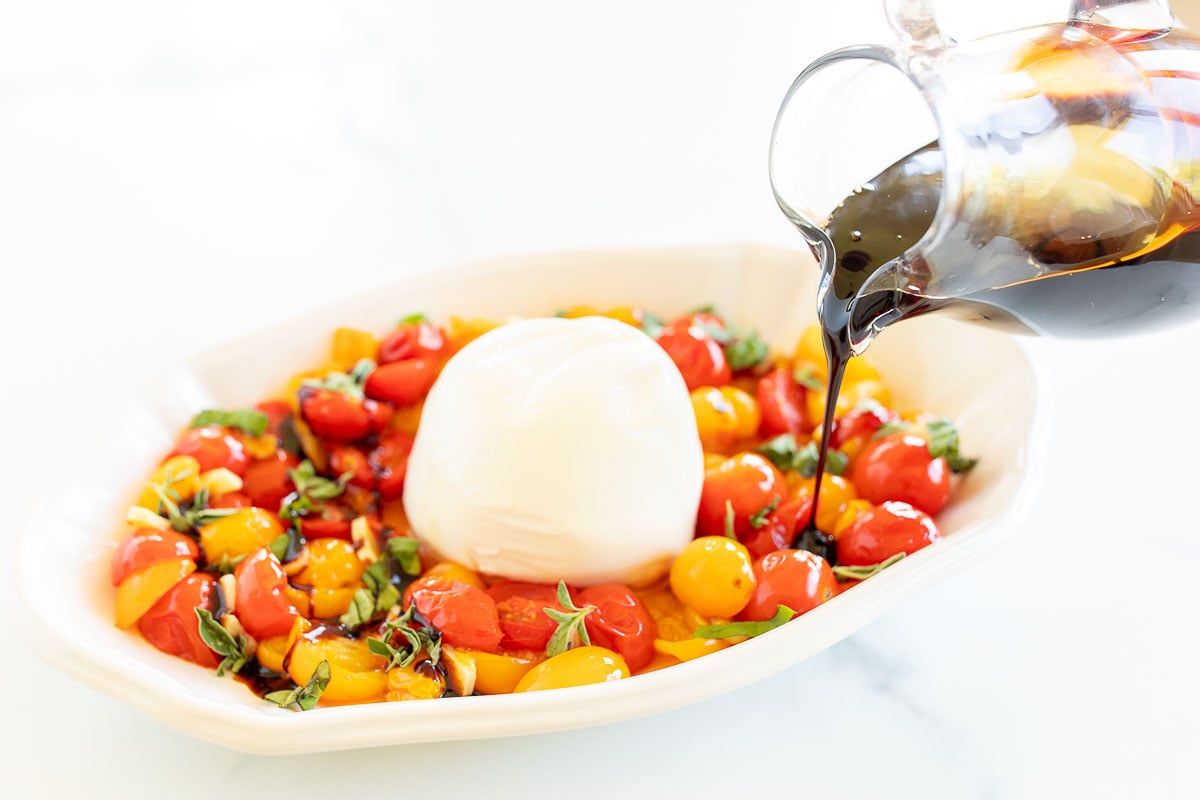 A small glass pitcher of balsamic glaze pouring over a dish of cherry tomatoes and mozzarella.