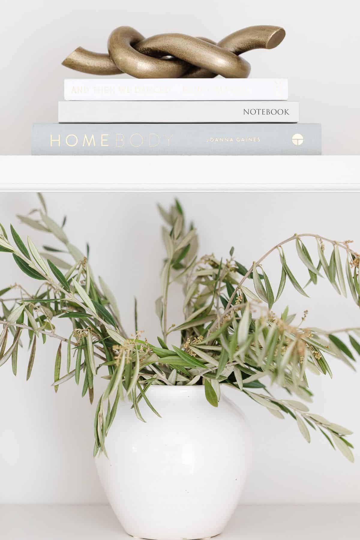 A white pottery vase full of greenery with books and a bookshelf decor above.