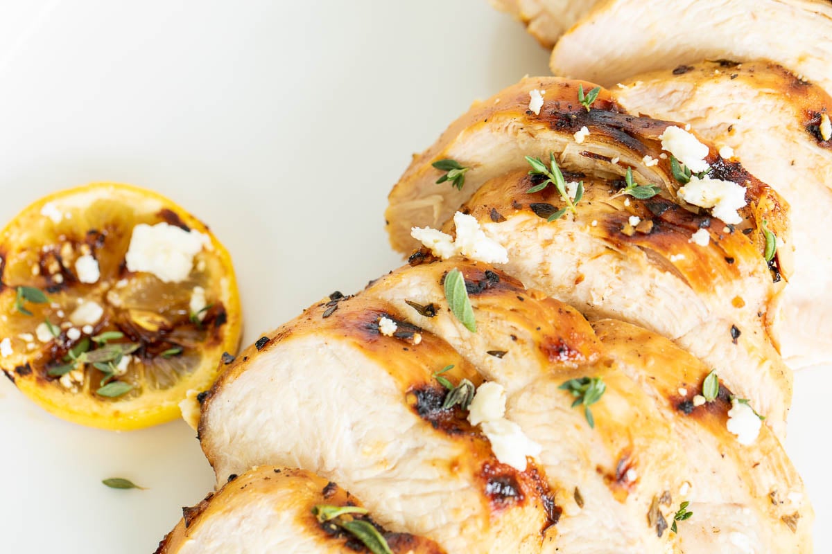 Slices of Greek marinated chicken on a white surface, grilled lemon to the side.