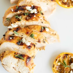 Slices of Greek marinated chicken on a white surface, grilled lemon to the side.