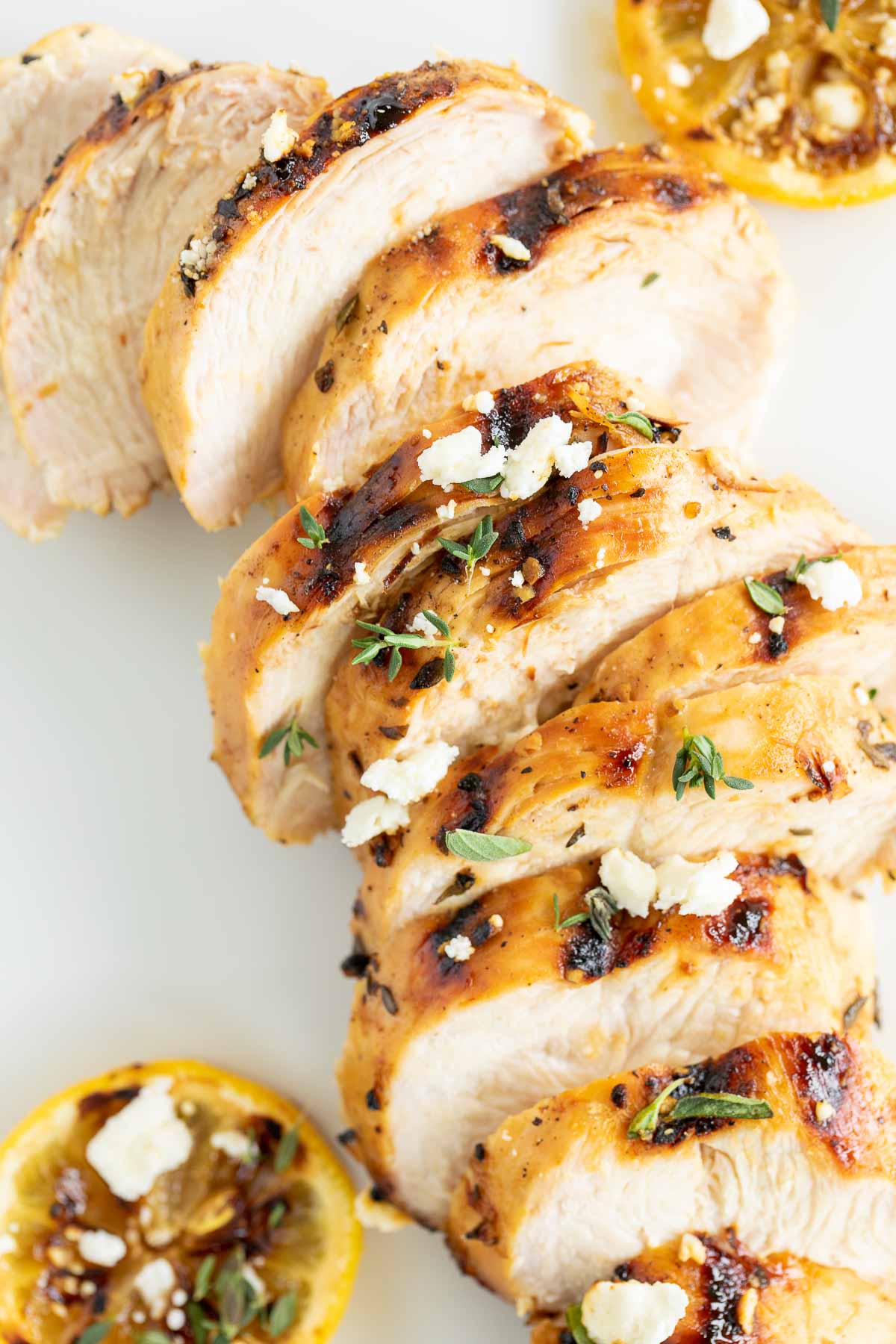 Slices of Greek marinated chicken on a white surface, grilled lemon to the side.