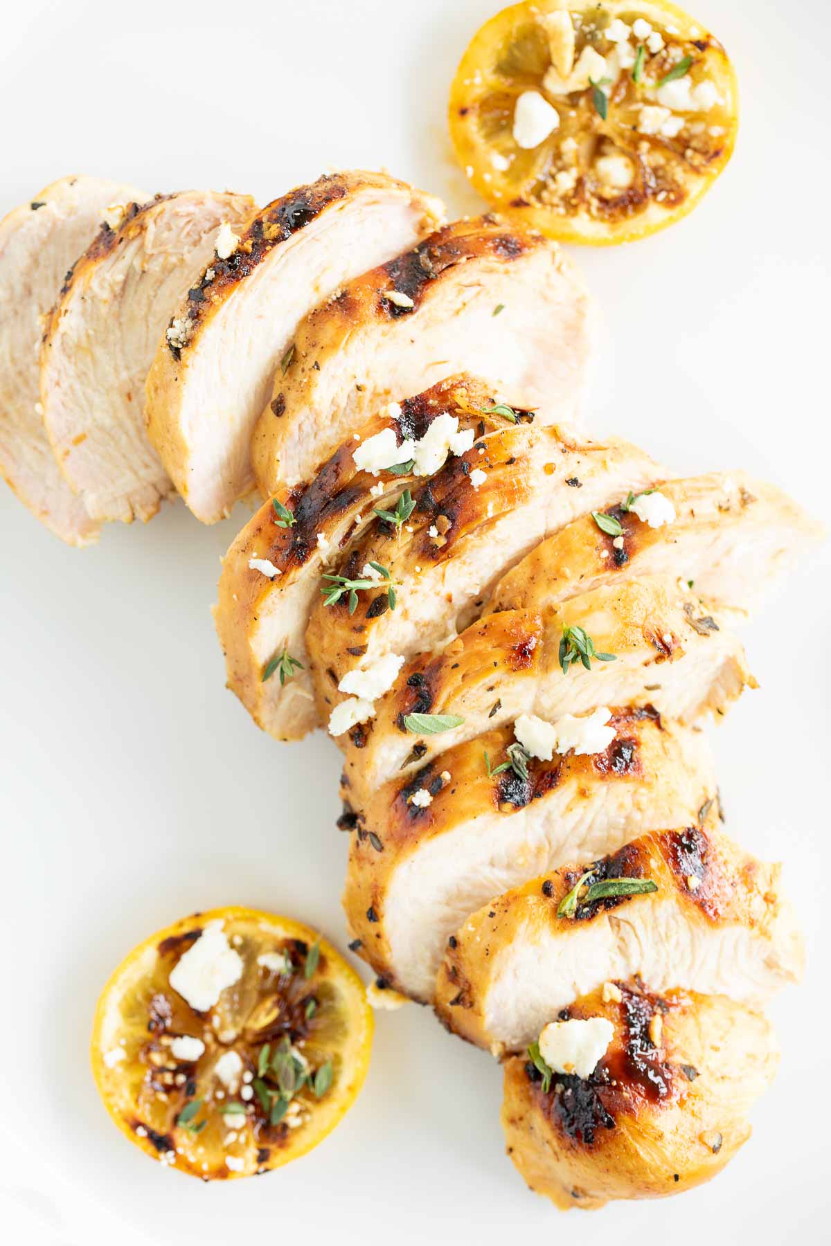 Slices of Greek marinated chicken on a white surface, grilled lemon to the side.