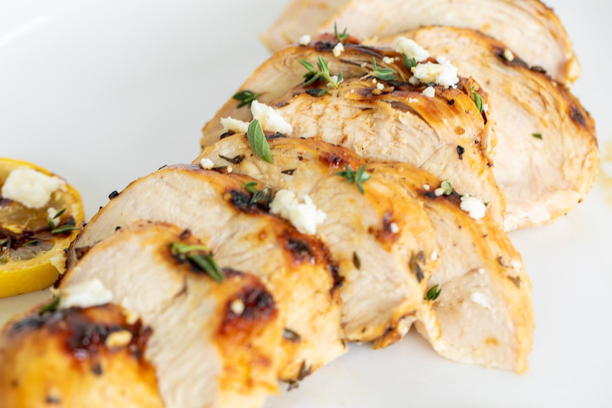 Slices of Greek marinated chicken on a white surface, grilled lemon to the side.