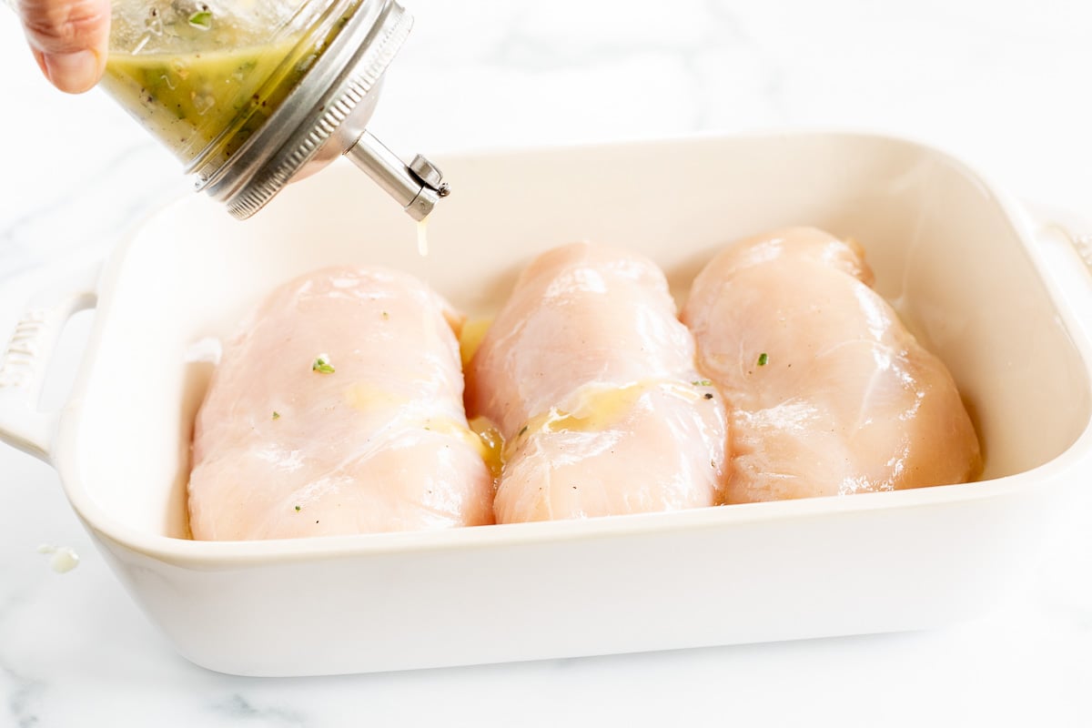 Greek chicken marinade in a jar, pouring over chicken breasts in a white dish.