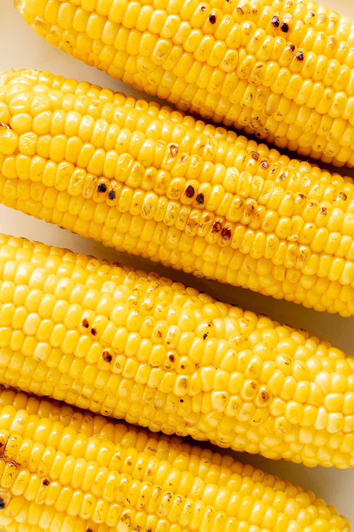 Four grilled corn on the cob arranged closely together on a white surface, showing golden yellow kernels with some charred spots.