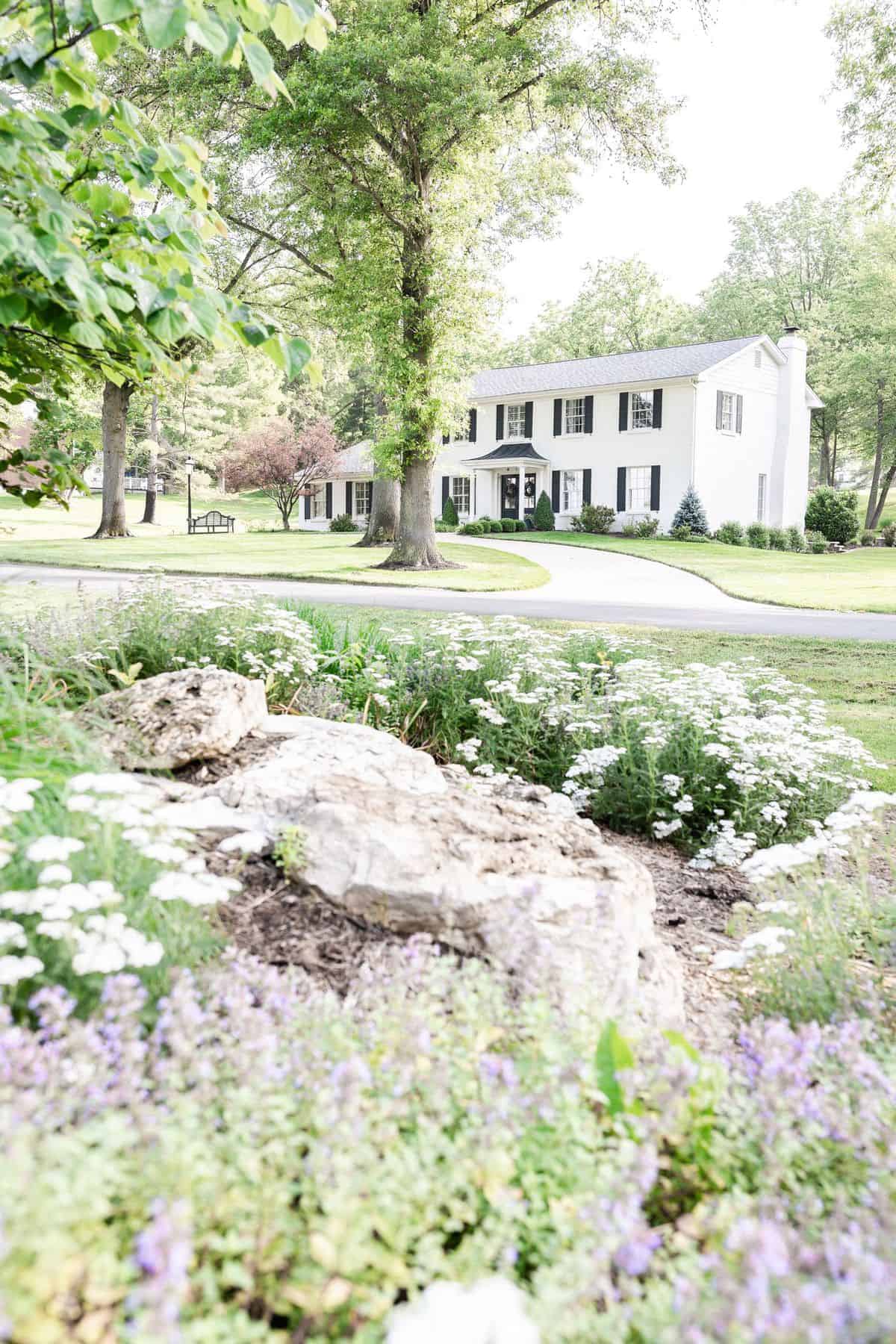 A white brick house sitting amongst green trees and flowers.