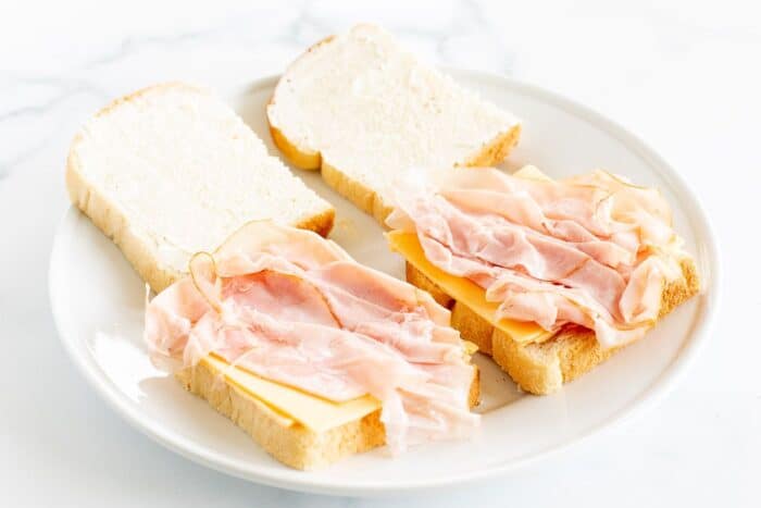 A white plate with sliced white bread topped with cheese and ham.