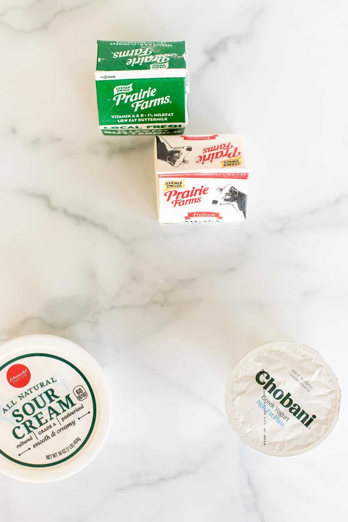 A small carton of buttermilk, a carton of whipping cream, a container of sour cream, and a small greek yogurt container all placed on a marble surface. 