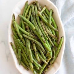 A white oval serving dish, filled with grilled green beans, blue linen towel to the side.